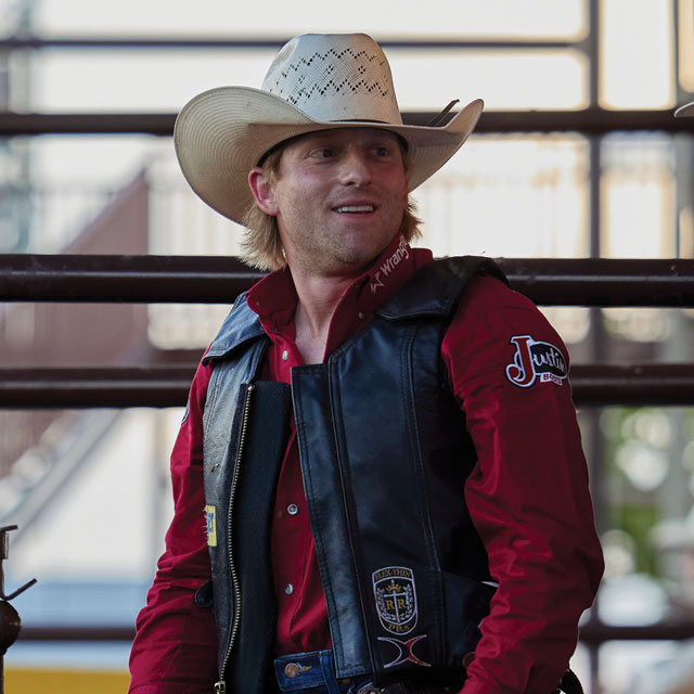 Brennon Eldred wearing a red shirt and a black vest looking away from the camera smiling. 